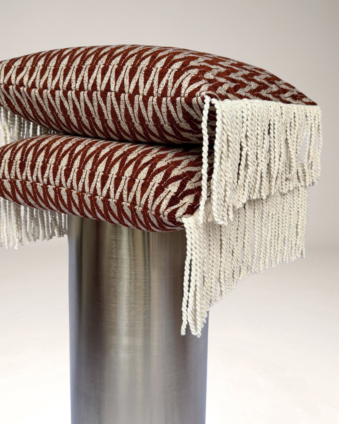 Hogaría sierkussen in terracotta en crème met grafisch geweven patroon en lange crèmekleurige franjes aan de zijkanten, op een chromen pilaar, luxe handgemaakt designkussen voor slaapkamer en woonkamer.