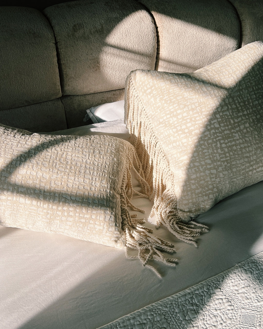 Crèmekleurige Hogaría-kussens met rijke textuur en franjes, gestyled op een zacht bed en belicht door natuurlijk zonlicht voor een serene, sfeervolle slaapkamerlook.