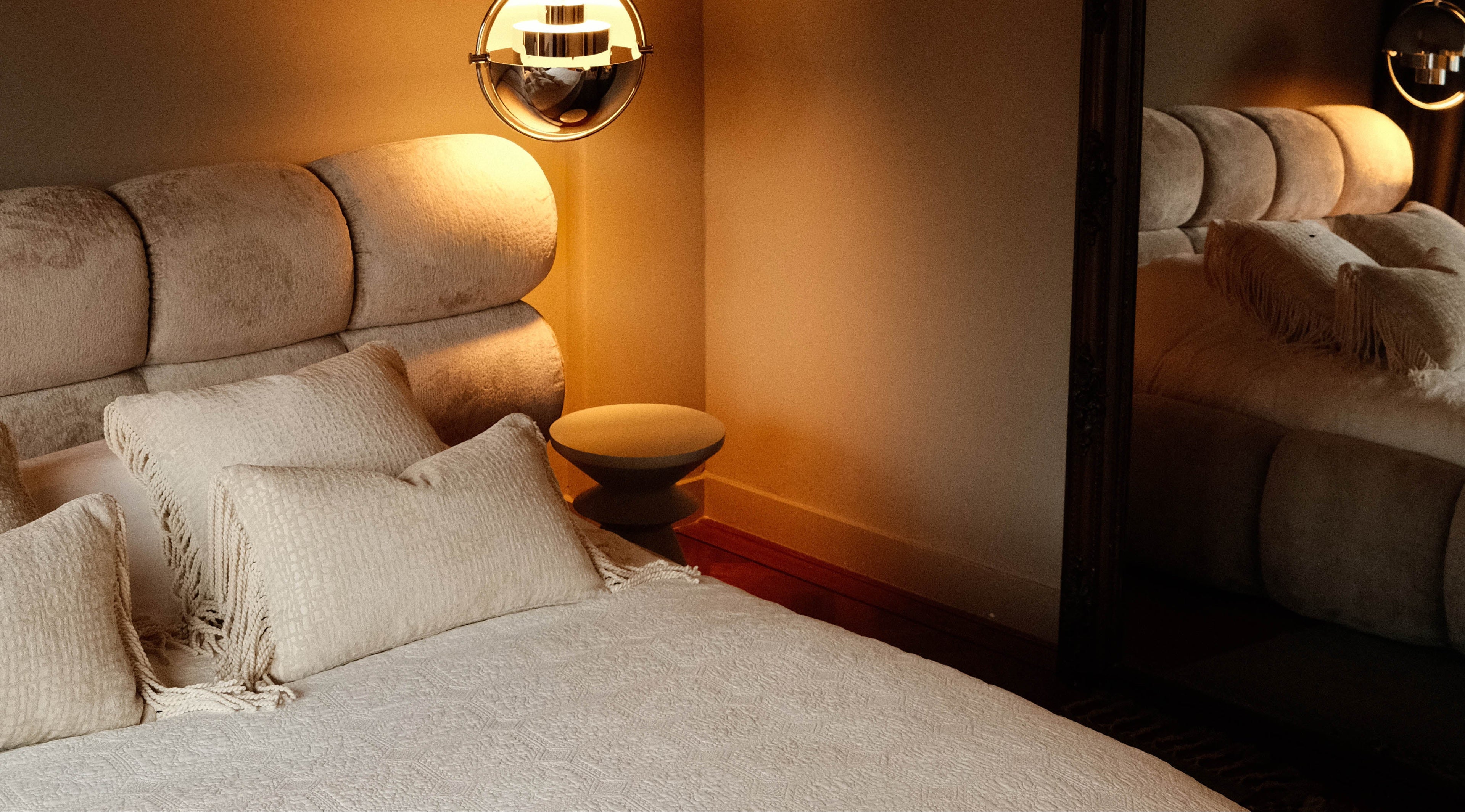 Sfeervol Hogaría slaapkamerinterieur met een beige hoofdbord, crème kussens met franjes en warme verlichting die in een grote spiegel weerspiegelt.